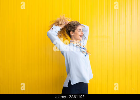 Lächelnde junge Frau ihr Haar binden vor gelbem Hintergrund Stockfoto