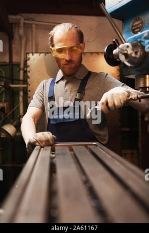 Bärtige junge Arbeiter in Schutzbrille arbeiten mit Metall auf speziellen Maschinen in der Fabrik Stockfoto