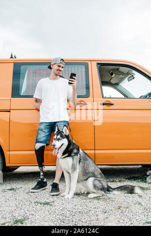 Junger Mann mit Hund tragen Beinprothese und über Handy an Wohnmobil Stockfoto