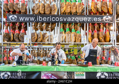 Aracena, Huelva, Spanien - Oktober 20, 2019: Metzger schneiden Scheiben eines spanischen Iberian Serrano Schinken mit einem Aussteller der ausgehärteten Iberico Schinken im Hinterg Stockfoto