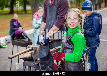 Vater und Kind sind im Handwerk tätig. Ein Schmied schmiedet eine Metall blank auf dem Amboss auf einer Messe in Anwesenheit von Zuschauern. Close-up. Stockfoto