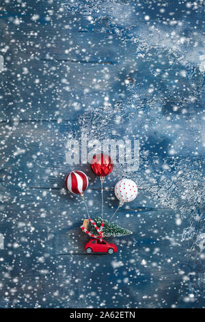 Miniatur Spielzeug rotes Auto mit Tannenbaum und Kugeln wie Luftballons auf dem verschneiten Hintergrund, Postkarte Konzept, getönt Stockfoto