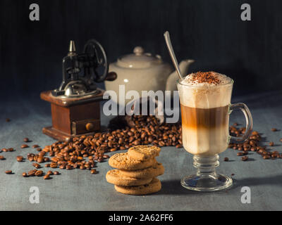 Hot Latte Macchiato in einem hohen Glas mit Schokolade Streusel auf der Oberseite. Im Hintergrund gibt es Kaffeebohnen verschüttet und eine Kaffeemühle. Stockfoto