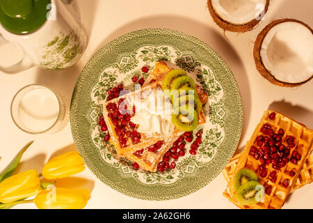 Hausgemachte frische belgische Waffeln mit Puderzucker bestreut und auf einer gestalteten Platte gelegt. Daneben ist ein Glas Milch, Tulpen im Krug Stockfoto