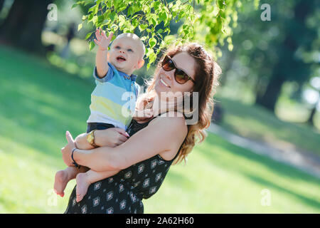 Die junge Mutter hält Ihr kleines Kind auf den Händen. Baby Boy trifft die Welt. Er schnappt sich die Zweige eines Baumes mit seinen Händen. Sie werden lachen. Stockfoto