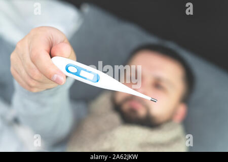 Krankheit. Zu Hause erfreuen. Ein junger Mann ist krank, ist zu Hause behandelt. Die Messung der Körpertemperatur. In den Händen eines Mannes ist ein elektronisches Thermometer. Mock Up Stockfoto