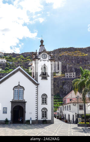 Ribeira Brava, Madeira, Portugal - Sep 9, 2019: Historische Römisch-katholische Kirche in der Altstadt. Palme auf den Platz. Bananenplantage auf einem felsigen Hang im Hintergrund. Stockfoto