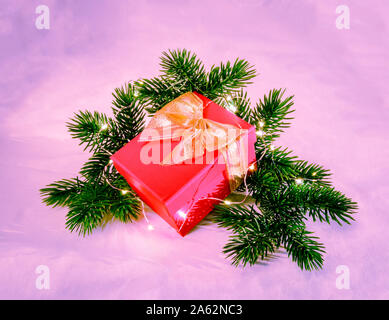 Rot Weihnachtsgeschenk Box mit Goldband und bowknot auf Fir Tree Branches mit funkelnden Lichtern geschmückt Girlande auf neon-lila Hintergrund Stockfoto