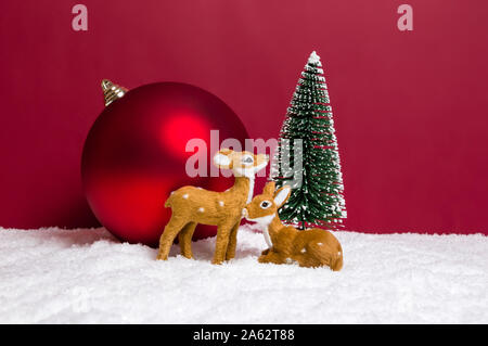 Weihnachten Thema mit zwei niedlichen Rentiere, Weihnachtsbäume und Große Christbaumkugel auf Schnee gegen den roten Hintergrund. Frohe Weihnachten Gruß und Miniatur Stockfoto