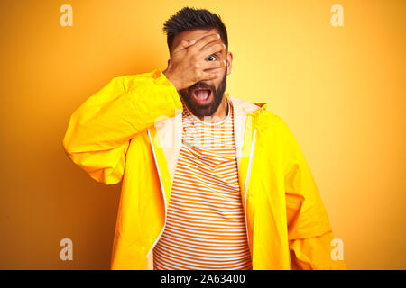 Junge indische Mann mit regenmantel stehend über isolierte gelben Hintergrund peeking im Schock für Gesicht und Augen mit der Hand, schauen durch die Finger Stockfoto