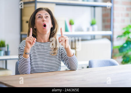 Mittleren Alters ältere Frau am Tisch sitzen zu Hause erstaunt und überrascht mit der Suche und zeigt mit Fingern und erhobenen Armen. Stockfoto