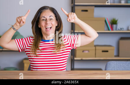 Mittleren Alters ältere Frau am Tisch zu Hause lächelnd erstaunt und überrascht und zeigt mit Fingern und erhobenen Arme sitzen. Stockfoto