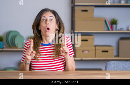 Mittleren Alters ältere Frau am Tisch sitzen zu Hause erstaunt und überrascht mit der Suche und zeigt mit Fingern und erhobenen Armen. Stockfoto