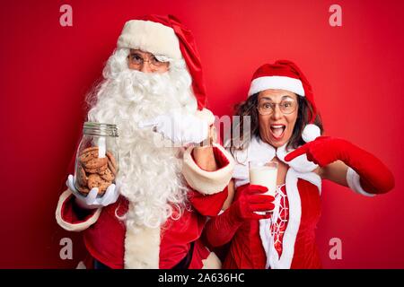 Paar Tragen santa Kostüm Holding jar von Cookies und Milch über isolierten roten Hintergrund sehr glücklich zeigte mit Hand und Finger Stockfoto