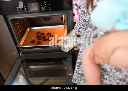 Die junge Mutter hält Ihr neugierigen kleinen Kind in ihren Armen helfen Ihr ein Dessert, das auf einem schönen Holztisch mit Puderzucker, Zimt vorbereiten, vanil Stockfoto