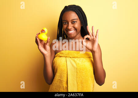 Afrikanische amerikanische Frau mit Handtuch holding Spielzeug Ente über isolierte gelben Hintergrund tun ok Zeichen mit den Fingern Dusche, ausgezeichnete Symbol Stockfoto