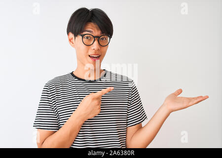 Chinesischer Mann mit Brille und Marine gestreiftes T-Shirt stehend über isoliert weißer Hintergrund erstaunt und lächelt in die Kamera während der Präsentation mit Ha Stockfoto