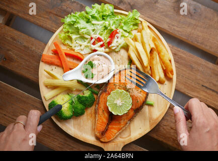 Lachs Steak serviert auf Holzplatte, Beilagen, gekochtes Gemüse, Pommes frites, Salat, Sauce und einem Stück Zitrone auf der Oberseite Stockfoto