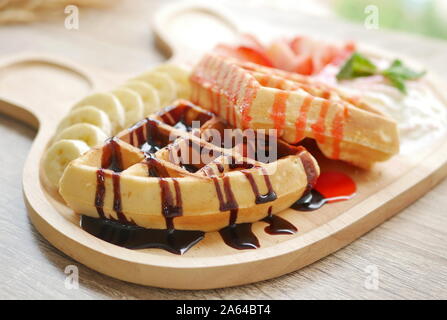 Leckere Waffel garniert mit Erdbeer Sirup und Schokoladensauce, Beilagen sind frische Erdbeere gehackt, Banane, Sahne und Eis serviert. Stockfoto