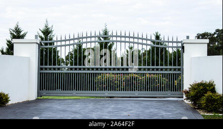 Graues metall Schmiedeeisen Auffahrt Eigenschaft Eingangstore in weißem Beton Ziegel Zaun, garten Bäume im Hintergrund Stockfoto