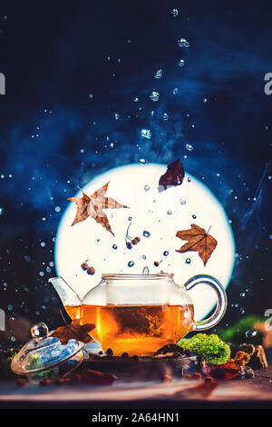 Herbst Tee Teekanne aus Glas mit strahlenden Vollmond, fallende Blätter und Wassertropfen. Kreative trinken Fotografie Konzept Stockfoto