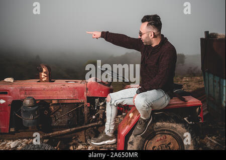 Armenischer Kaukasischer Mann sitzt auf dem alten gebrochenen roten Traktor in den Bergen während des nebligen Tages. Die interessanten verlassenen Dinge während der Reise zu finden Stockfoto