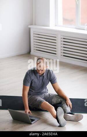 Junge behinderte Menschen sitzen auf dem Boden auf der Trainingsmatte und online Sport Training auf Laptop im Zimmer Stockfoto