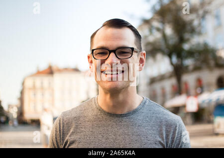 Die Hälfte Körper geschossen des jungen Mannes in einer Straße, einer alten Stadt in Europa, die in der Kamera mit braunen Augen und eyesglasses Stockfoto