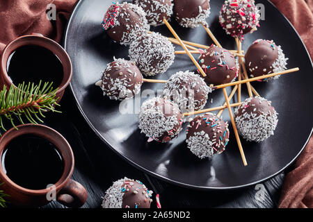 Nahaufnahme der festlichen Schokoladenkuchen pops bestreut mit zerquetschten Bonbons und Kokosraspeln auf einer schwarzen Platte. Tassen Kaffee, tannenzweigen, braun Tuch auf Stockfoto