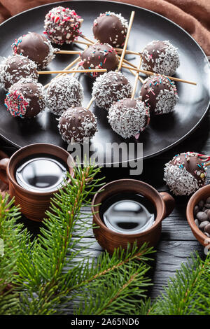Nahaufnahme der festlichen Schokoladenkuchen pops bestreut mit zerquetschten Bonbons und Kokosraspeln auf einer schwarzen Platte. Tassen Kaffee, tannenzweigen, braun Tuch auf Stockfoto