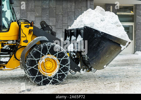 Big loader Maschine mit Stahl Ketten aus Metall entfernen von großen Schnee Haufen vom city street bei alpine Bergwelt im Winter. Starker Schneefall Nachwirkungen Stockfoto