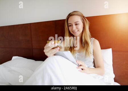 Frau Kauf von Waren online zufrieden mit Debit Kreditkarte und Tablet Verlegung in weißen Bett zu Hause mit Sonnenschein Stockfoto
