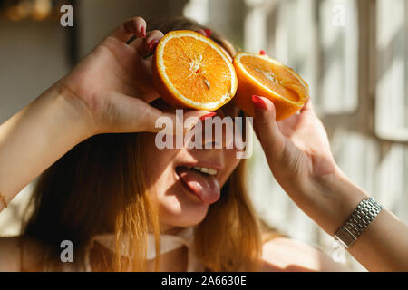 Schönheit Ginger Girl nimmt saftigen Orangen. Schöne freudige Teen Girl Holding Orangenscheiben und mit Zunge, lachen Emotionen. Stockfoto