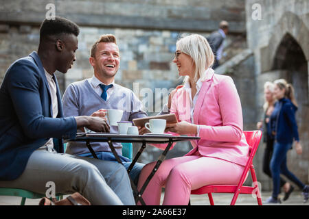 Zwei Geschäftsleute und eine Geschäftsfrau, die an einem Tisch sitzen in einer Stadt in einem heißen Getränk und etwas auf einem digitalen tablet Lachen. Ein Mann Stockfoto