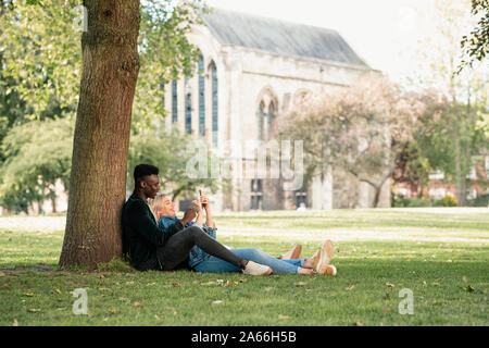 Ein Mann und eine Frau sitzen in einem Park gegen einen Baum. Die Frau sitzt zwischen den Beinen der Mann, während ihm etwas auf ihr Mobiltelefon. Stockfoto