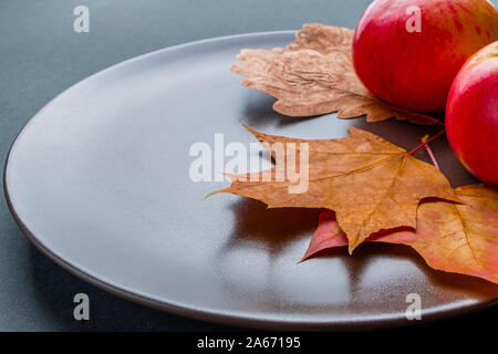 Blick auf rote Äpfel und bunte Blätter im Herbst liegen auf einem braunen Schüssel, auf einem dunkelgrauen Hintergrund. Schrägansicht mit Platz für Text. Stockfoto