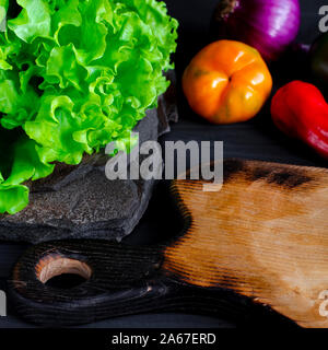 Rote, gelbe, grüne Paprika, Chili, Zwiebeln und grünem Salat liegen auf einem schwarzen Holztisch, neben einem Schneidbrett auf dem grünen Pfeffer und ein Stockfoto