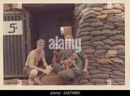 Drei amerikanische Soldaten, eine multirassische Gruppe bestehend aus zwei kaukasischen Männern und einem afroamerikanischen Mann, posieren mit einem Welpen und Dosen Carling Black Label Beer vor Sandsäcken auf einer Befestigung im Dschungel in Vietnam während des Vietnamkriegs, 1975. () Stockfoto