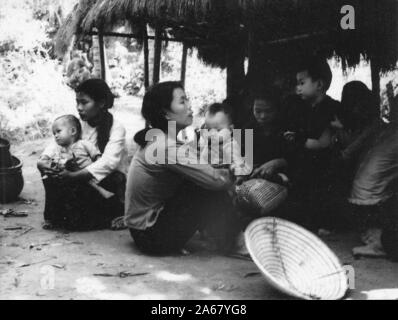 An einem sonnigen Tag, Vietnam 1965, sitzen zivile vietnamesische Frauen und Kinder im Schatten der Strohdachhütte zusammen. () Stockfoto