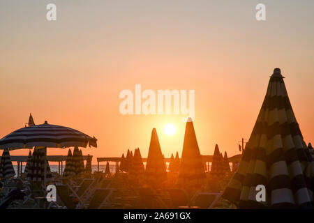 Hintergrundbeleuchtung Blick auf einen Strand an der Küste der Versilia in der Toskana mit Reihen von Sonnenschirmen und orange Himmel bei Sonnenuntergang im Sommer, Lido di Camaiore, Italien Stockfoto