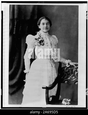 Junge afrikanische amerikanische Frau, drei-viertel länge Portrait, stehend, mit leicht nach rechts, mit der linken Hand auf der Rückseite des Stuhls Stockfoto