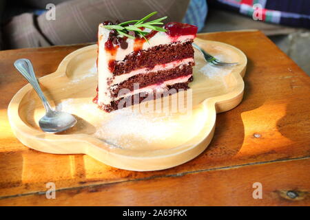 Red Velvet Cake mit Erdbeer Smoothie und Sirup, süße Speisen mit Löffel und Gabel auf einer Holzplatte mit braunen Tisch im Hintergrund Stockfoto