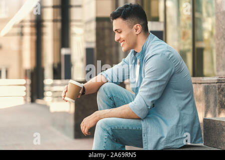 Freizeitaktivitäten im Freien. Stilvolle Kerl sitzt auf der Treppe auf der Straße trinken heißen Kaffee Lachen glücklich Seitenansicht Stockfoto