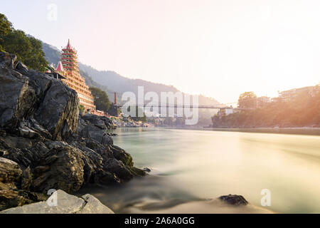 Atemberaubende Aussicht auf die Ufer des Flusses Ganga mit dem Lakshman Jhula Brücke und Tera Manzil Tempel (Trimbakeshwar) bei einem schönen Sonnenuntergang. Stockfoto