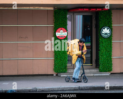 Moskau, Russland - Oktober 19, 2019: Asiatische junge Mann, ein Yandex-Eda Kurier in einer gelben Jacke mit Unternehmenskennzeichen, steht auf einer Straße der Stadt. Krieg Stockfoto
