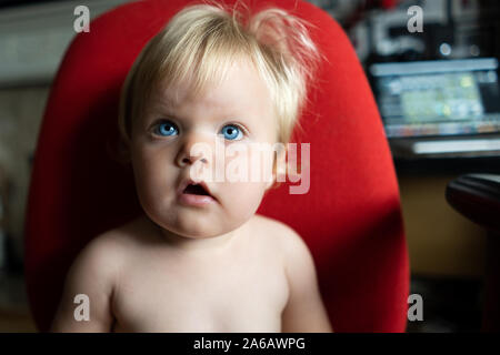 Ein hübsches kleines Mädchen mit blondem Haar und blauen Augen sitzen auf einem roten Bürostuhl, etwa 18 Monate alt Stockfoto