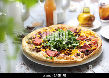 Italienische Pizza mit Mozzarella Käse, Salami, Mais und Rucola. Auf einer Platte und einem weißen Tisch serviert. Stockfoto