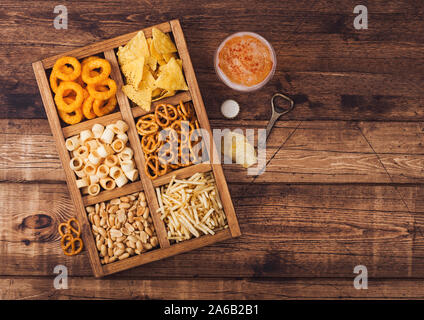 Glas Handwerk Lagerbier und Opener mit Box von Snacks auf Holz Hintergrund. Brezel, salzig Kartoffelstifte, Erdnüsse, Zwiebelringen mit Nachos in Vintage b Stockfoto