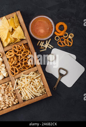 Glas Handwerk Lagerbier und Opener mit Box von Snacks auf dunklem Hintergrund. Brezel, salzig Kartoffelstifte, Erdnüsse, Zwiebelringen mit Nachos in Vintage b Stockfoto