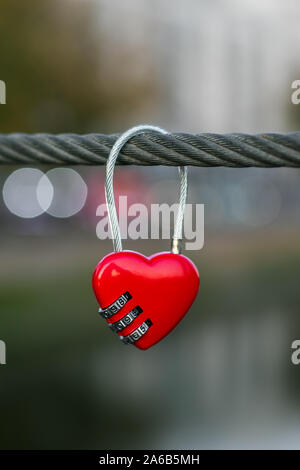 Rotes Vorhängeschloss an einem Seil, Bokeh des abends die Lichter der Stadt. Symbol der Liebe. Romantische Konzept. Metal Heart an einem Kabel auf der Brücke über den Fluss. Herz hängen an kleiden Stockfoto